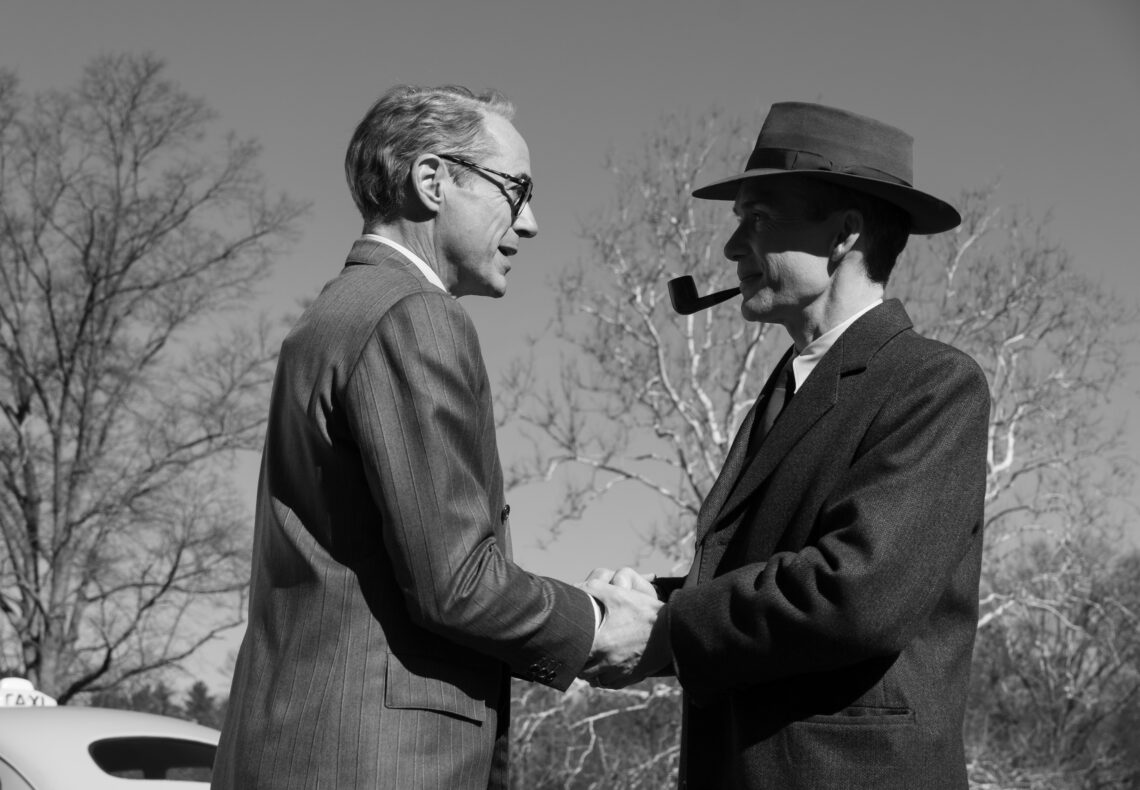 Schwarz-Weiss Screenshot aus dem Film Oppenheimer. Links steht Lewis Strauss (Robert Downey Jr.) und rechts steht Robert Oppenheimer (Cilian Murphy), sie sehen sich an und schütteln sich die Hände
