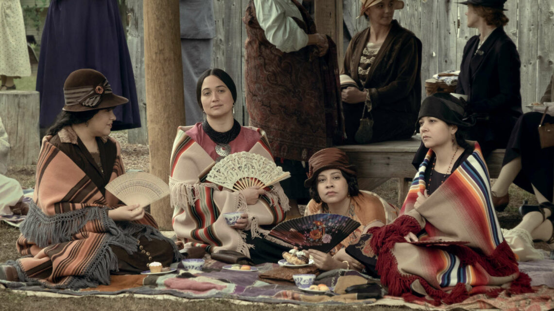 Szene aus dem Film. Vier junge indigene Frauen mit elegangen Fächern sitzen auf einer Decke und beobachten das Geschehen.