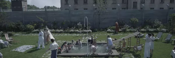 Szene aus dem Film The Zone of interest. Im Vordergrund ein großer Garten mit Steinwegen und einem kleinen Swimmingpool in der Mitte in dem Kinder spielen. Im Hintergrund eine Mauer und dahinter das Konzentrationslager Auschwitz.