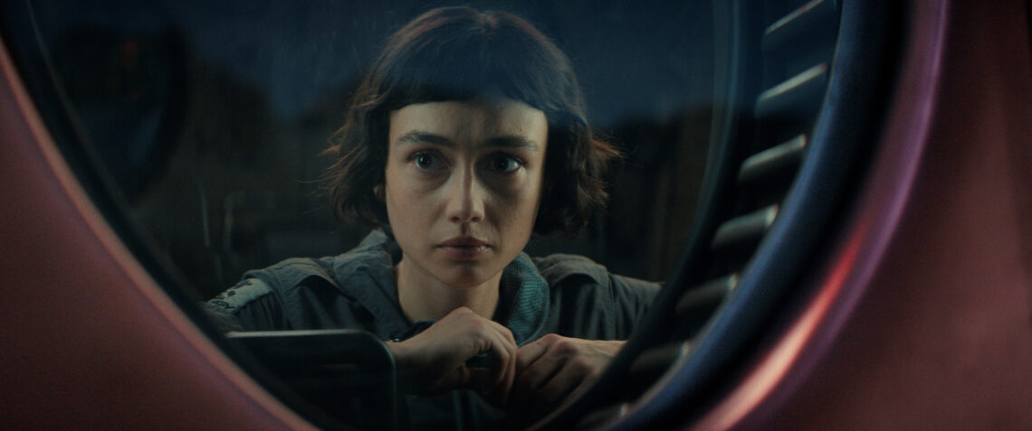 a young white woman with bowl cut hair looking serious outside a round window
