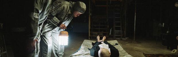 Szene aus dem Film Bugonia: Zwei Männer in silbernen Schutzanzügen beugen sich über eine Frau in Business Kleidung, die auf einer Liege in einem dunklen Keller liegt. Copyright Universal Pictures 2025